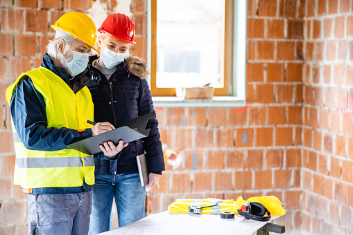 Senior male construction site engineer and female client in protective face masks discussing building blueprint plans on the new house