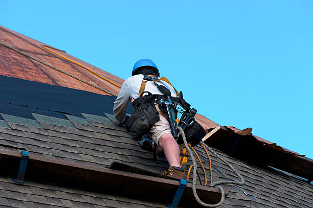 A construction worker with tools and safety equipment scales a large roof while nailing on ashpalt shingles.